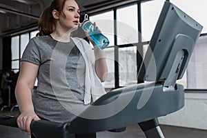 Curvy girl drinking from water bottle while running on treadmill