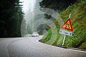 Curvy mountain road with slippery route sign and blurred white car in the background