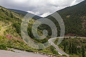 Curvy paved road winding through a forested mountain range under a rainy cloudscape