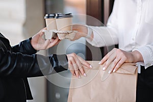 Customer in face mask stand outdoors and take package with coffee and food from cashier in mask at cafe. Take away food