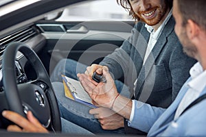 A customer is sitting in the car at auto shop with a salesperson and receiving the keys of a vehicle he bought. Car, shop, buying