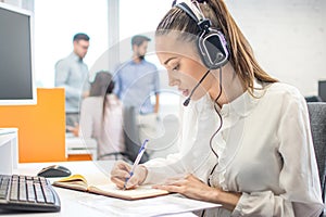Customer support operator woman talking to a client and writing notes to notebook in call center.