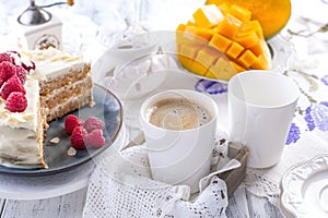 Cut the cake with white cream, for breakfast. A mango fruit. White background, tablecloth with lace, a cup of fragrant black