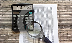 Cut cost, the text is written on the display of the word calculator. Documents and a magnifying glass are on a wooden table