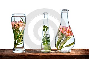 Cut flowers and tropical plants in a glass of water on a barn wood table on white background