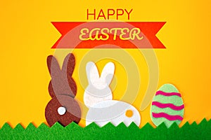 Cut out of felt applications of easter egg and white and brown rabbits on the grass. Yellow background. Flat lay. Easter holiday