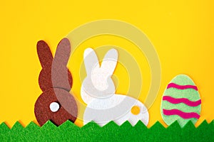 Cut out of felt applications of easter egg and white and brown rabbits on the grass. Yellow background. Rear view. Flat lay.