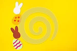 Cut out of felt applications of easter multicolored eggs and silhouette of white and brown rabbits. Yellow background. Flat lay.