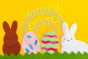 Cut out of felt applications of two eggs and white and brown rabbits on the grass. Yellow background. Flat lay. Easter holiday