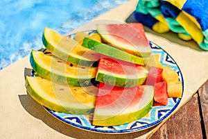 Cut slices and cubes of red and yellow watermelon on a patterned ceramic plate on the edge of a swimming pool. Colorful striped po