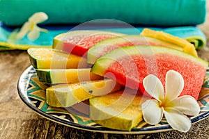 Cut slices of red and yellow watermelon decorated with a Plumeria or Frangipani flower on a colorful patterned ceramic plate