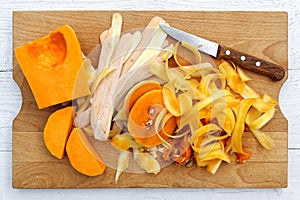 Cut up butternut squash with peels and knife on brown wood chopping board isolated on white from above.