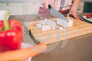 Cut view of female`s hands cutting white chees into pieces on desk. She uses knife. There are carrot and pepper on plate.