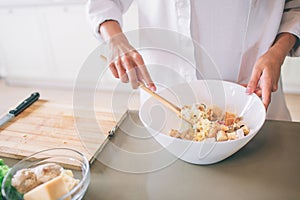 Cut view of girl`s hands mixing salad in white bowl. She is using spoon for that. Girl wears white shirt. She cooks.