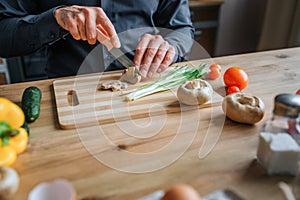 Cut view of man` hands cutting mushrooms with knife. He work with both hands. Man stand in kitchen at table.