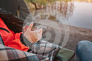 Cut view of oyung woman`s hands holding cup with hot drink. She sits at the edge of water and covered with blanket