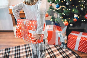 Cut view of small girl standing and holding red box behind back. It is present. There are another gifts under Christmas