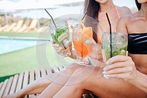 Cut view of three beautiful women sit on sunbeds and hold cocktails in hands. There are two green and one orange. Models