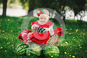 Cute 10 months baby girl on picnic on grass with watermelons. smiling child in red dress and bow on head. Concept of