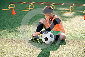 Cute 13-aged football player on artificial green covering of outdoors sport field and tying the shoelace on his boots.