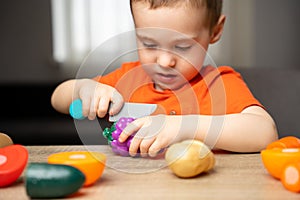 A cute 3 year toddler boy plays with toy fruits or vegetables and sitting at the table.