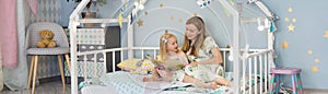 Cute 3 years old girl and her young mother are reading a book and smiling while sitting in decorated house bed at