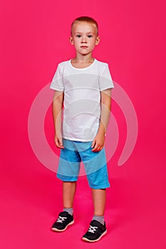 Cute 5-year-old boy in a white T-shirt and blue full-length shorts on a pink isolated background. Vertical