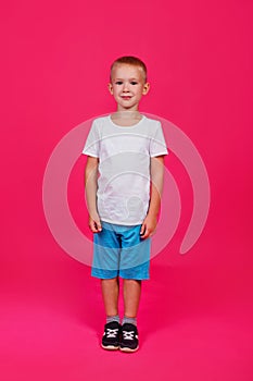 Cute 5-year-old boy in a white T-shirt and blue full-length shorts on a pink isolated background. Vertical