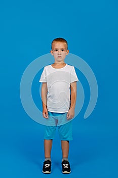 A cute 5-year-old boy in a white t-shirt and blue shorts. Full growth. Blue background. Vertical