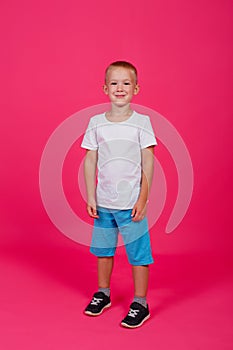 A cute 5-year-old boy in a white t-shirt and blue shorts. Full growth. Pink background. Vertical
