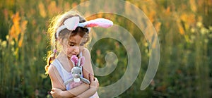 Cute 5-year-old girl with rabbit ears gently hugs a toy rabbit in nature in a blooming field in summer with golden sunlight.