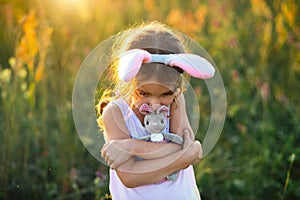 Cute 5-year-old girl with rabbit ears gently hugs a toy rabbit in nature in a blooming field in summer with golden sunlight.