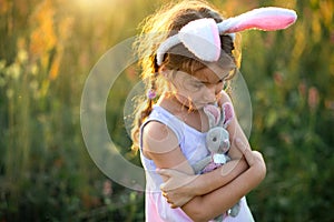 Cute 5-year-old girl with rabbit ears gently hugs a toy rabbit in nature in a blooming field in summer with golden sunlight.