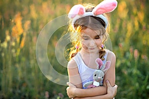 Cute 5-year-old girl with rabbit ears gently hugs a toy rabbit in nature in a blooming field in summer with golden sunlight.