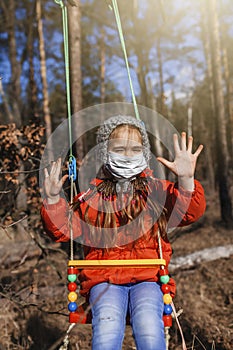 A cute 6-7 years old girl in red coat wearing respirator mask flying on the swing alone in the forest