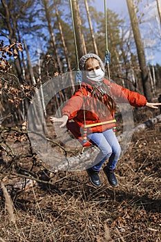 A cute 6-7 years old girl in red coat wearing respirator mask flying on the swing alone in the forest