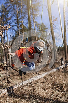 A cute 6-7 years old girl in red coat wearing respirator mask flying on the swing alone in the forest