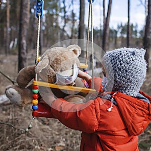 A cute 6-7 years old girl in red coat wearing respirator mask on her teddy bear toy on the swing alone