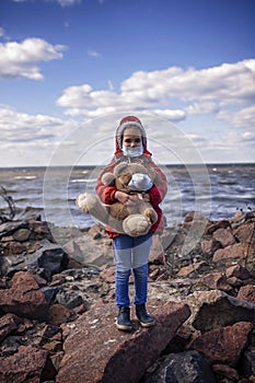 A cute 6-7 years old girl in red coat wearing respirator mask playing with toy teddy bear alone