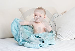 Cute 9 months old baby boy covered in blue towel sitting on bed after bathing