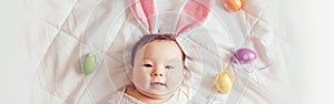 Cute adorable Asian baby wearing pink Easter bunny ears. Infant kid lying on bed with coloredl Easter eggs. Funny child