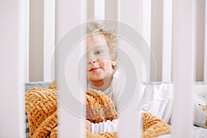 Cute adorable baby boy toddler lying in crib at kids nursery room at home. Funny charming baby boy with curly blond hair and blue