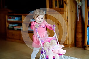 Cute adorable baby girl make first steps and push with doll carriage. Beautiful toddler child pushing stroller with toy