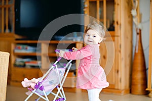 Cute adorable baby girl make first steps and push with doll carriage. Beautiful toddler child pushing stroller with toy