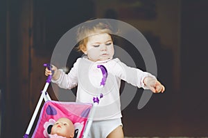 Cute adorable baby girl making first steps with doll carriage. Beautiful toddler child pushing stroller with toy at home