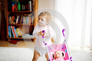 Cute adorable baby girl making first steps with doll carriage. Beautiful toddler child pushing stroller with toy at home