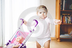 Cute adorable baby girl making first steps with doll carriage. Beautiful toddler child pushing stroller with toy at home