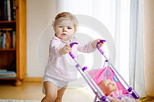 Cute adorable baby girl making first steps with doll carriage. Beautiful toddler child pushing stroller with toy at home