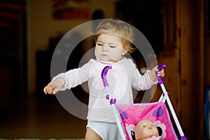 Cute adorable baby girl making first steps with doll carriage. Beautiful toddler child pushing stroller with toy at home