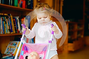 Cute adorable baby girl making first steps with doll carriage. Beautiful toddler child pushing stroller with toy at home